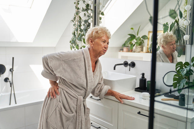 Senior person experiencing back pain in a bright, modern bathroom not featuring an adjustable-height sink set at a comfortable seated level to speed up a morning routine.